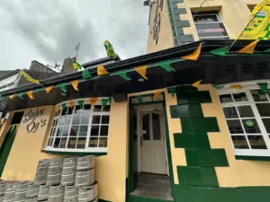Sean Og's pub in Ballyshannon with cream and green painted walls, decorated with green and yellow bunting and flags. Metal beer kegs are stacked by the entrance, under a cloudy sky—a classic Irish bar atmosphere.