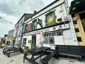 A street view of O'Donnell's Bar, a classic Irish pub in Ireland, features white walls with dark green trim, outdoor benches, hanging flags, and cloudy skies. The bar’s name is displayed above the entrance.