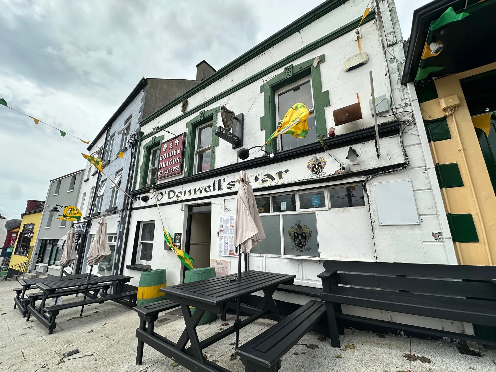 A street view of O'Donnell's Bar, a classic Irish pub in Ireland, features white walls with dark green trim, outdoor benches, hanging flags, and cloudy skies. The bar’s name is displayed above the entrance.