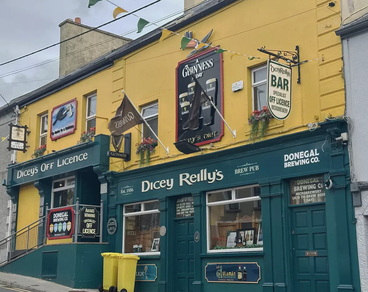 A vibrant yellow and green pub and off-licence building, "Dicey Reilly’s," with Guinness signs, flower boxes, and Donegal Brewing Co. branding, stands on a street in Ballyshannon, Ireland.