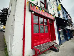A red storefront with a sign reading “Finn McCool’s” on a white building hints at a cozy Ireland pub. Donegal flags hang above, and a wooden bench sits in front on the sidewalk.