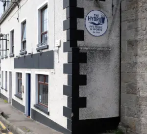 A white building with black trim, resembling a classic Ireland pub, displays a round sign reading "The Hydro – Live Music & Sports, Est. 2025." The bar features several windows and sits along a narrow street.
