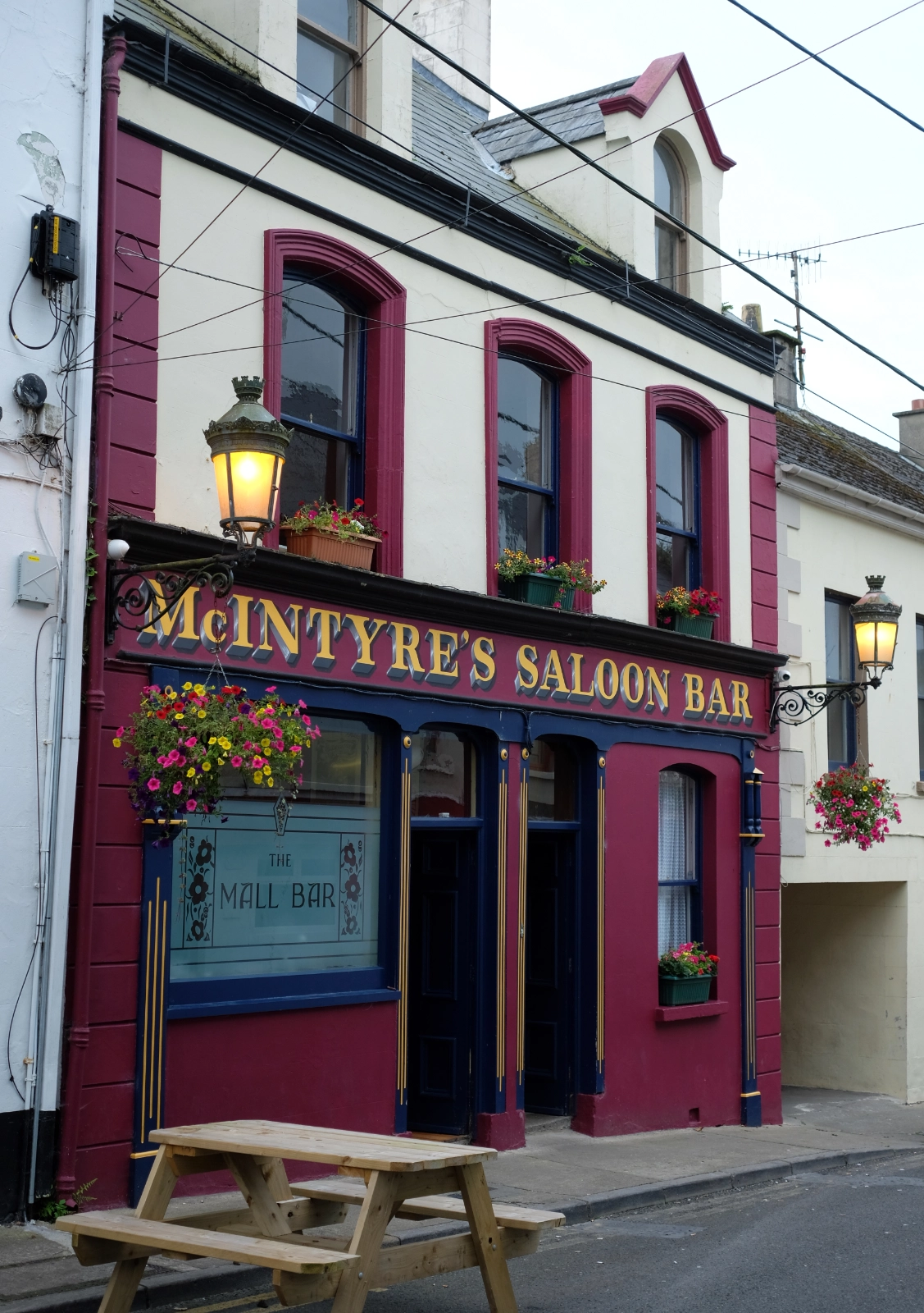 The image shows the exterior of McIntyre's Saloon Bar, a colorful pub in Ballyshannon, Ireland, with maroon and cream trim, hanging flower baskets, classic lanterns, and a wooden picnic bench outside on the street.