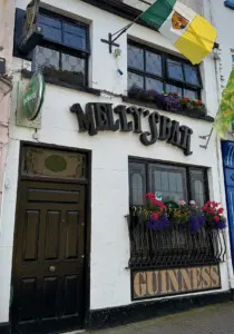 The front of Melly’s Bar in Ballyshannon features black signage, colorful flowers in window boxes, Guinness and Heineken signs, and Irish flags hanging above the entrance.
