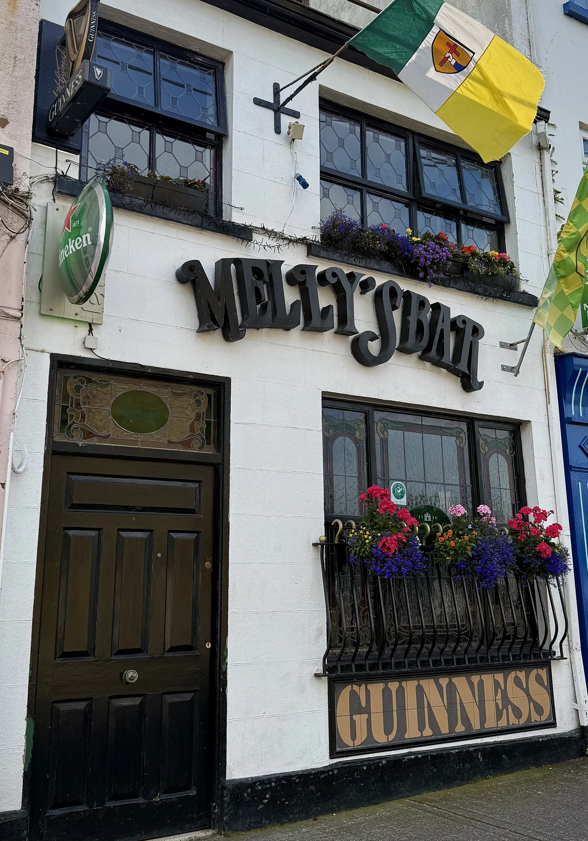 The front of Melly’s Bar in Ballyshannon features black signage, colorful flowers in window boxes, Guinness and Heineken signs, and Irish flags hanging above the entrance.