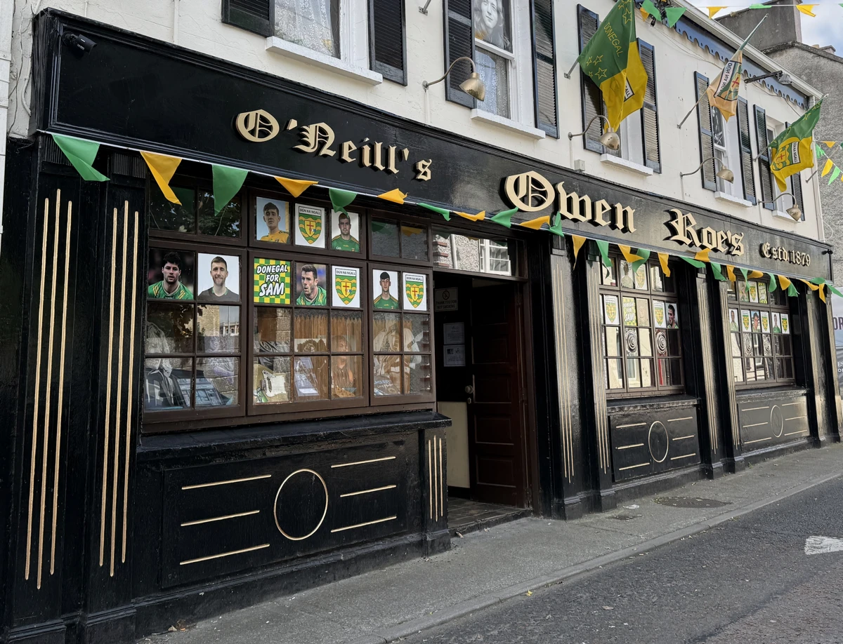 A traditional Irish pub in Ballyshannon, Owen Roe's displays yellow and green Donegal flags and sports posters in its windows. The black facade features gold trim and large windows facing the street.